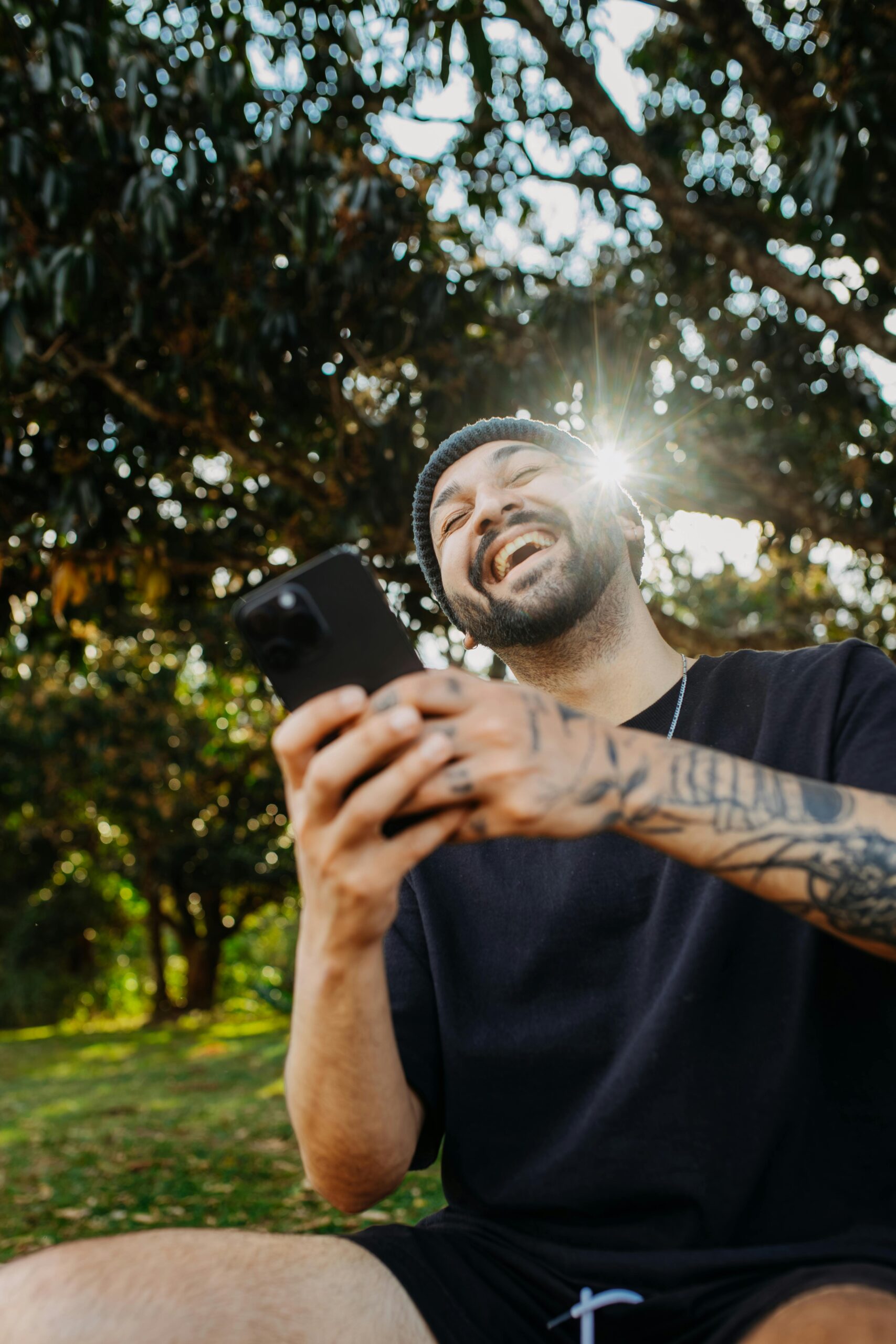 A cheerful man with tattoos laughing while holding a smartphone outdoors in a sunny park.